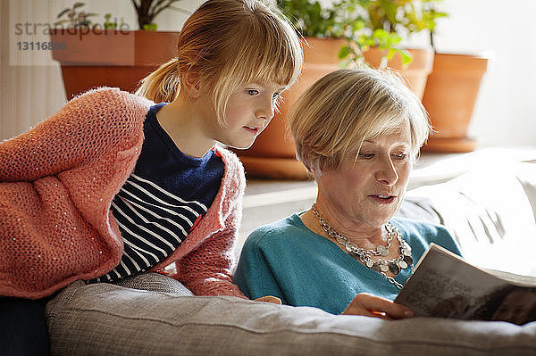 Mädchen hört Großmutter beim Zeitschriftenlesen zu  während sie zu Hause auf dem Sofa sitzt