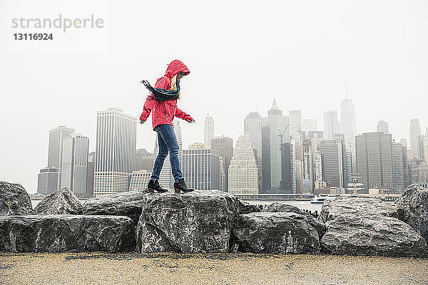 Seitenansicht einer Frau in rotem Regenmantel  die auf Felsen gegen Gebäude in der Stadt läuft