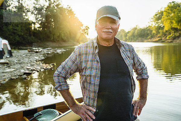 Porträt eines älteren Mannes mit Händen auf der Hüfte  der bei Sonnenuntergang am Ufer des Sees am Boot steht