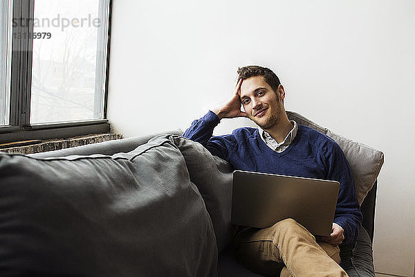 Lächelnder Mann benutzt Laptop  während er zu Hause auf dem Sofa sitzt