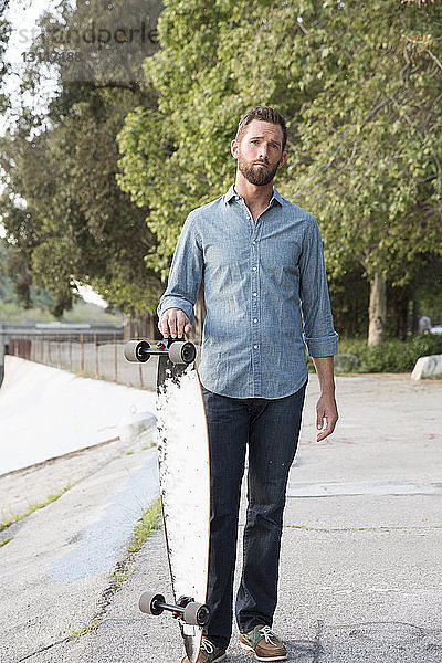 Porträt eines gut aussehenden Mannes mit Skateboard in der Hand an Bäumen stehend