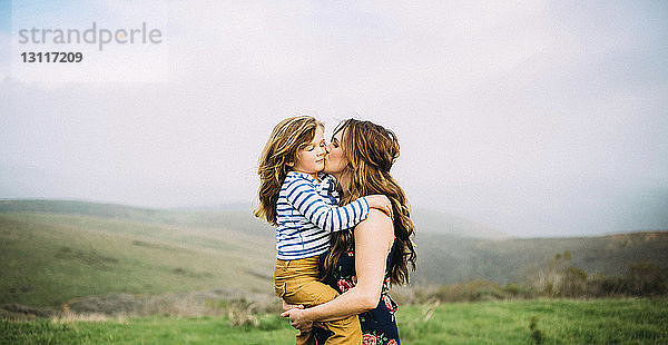 Liebende Frau küsst Tochter  während sie auf Feld gegen Himmel steht