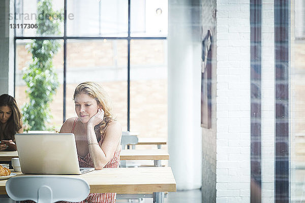 Frau mit Hand am Kinn  die einen Laptop benutzt  während sie mit einem Freund im Hintergrund durch ein Fenster in einem Café sitzt