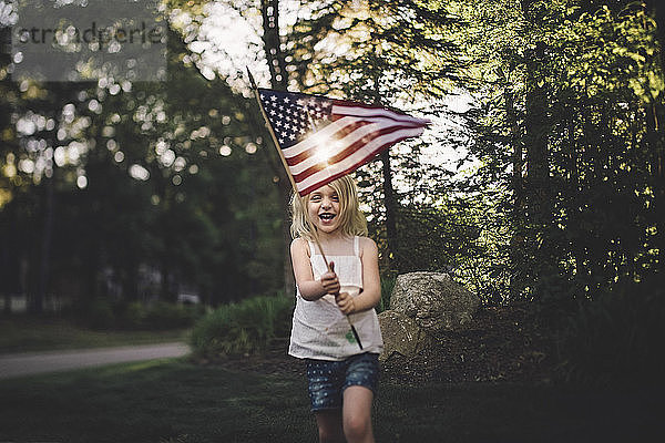 Fröhliches Mädchen mit amerikanischer Flagge läuft bei Sonnenschein im Hof