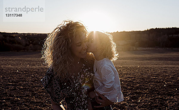 Liebende Tochter küsst fröhliche Mutter  während sie von ihr bei Sonnenuntergang auf dem Feld getragen wird
