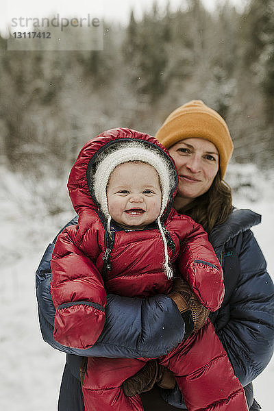 Porträt einer Mutter  die eine Tochter trägt  während sie im Winter im Wald steht