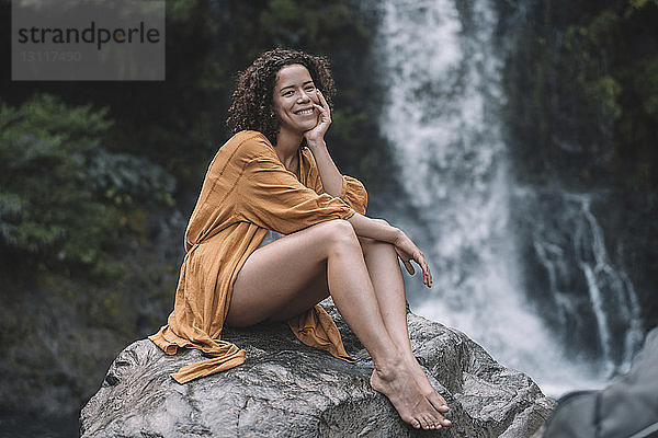 Porträt einer lächelnden Frau  die auf einem Felsen vor einem Wasserfall im Wald sitzt