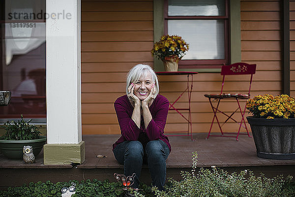Porträt einer fröhlichen älteren Frau mit Händen am Kinn auf der Veranda sitzend