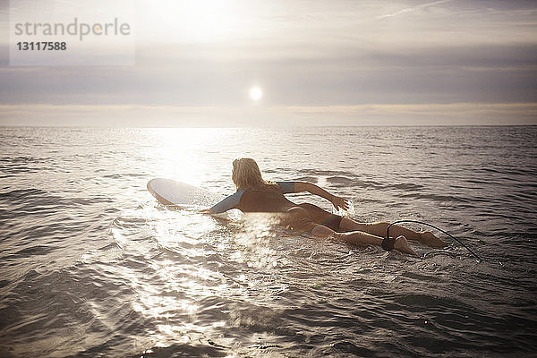 Rückansicht einer Surferin  die bei Sonnenuntergang auf einem Surfbrett im Meer liegt