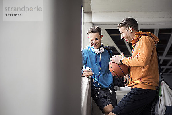 Glückliche männliche Freunde mit Basketball mit Smartphone auf dem Parkplatz