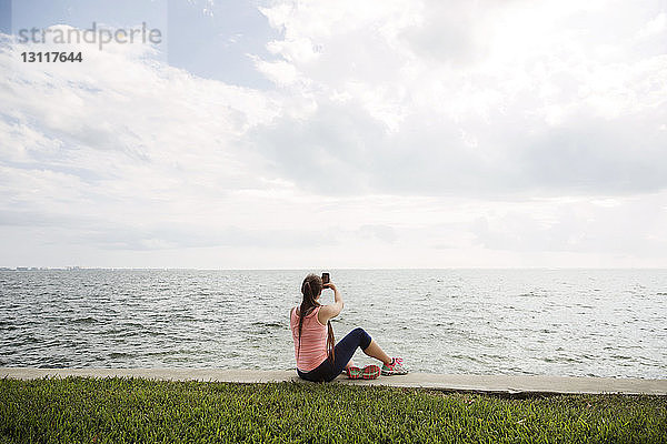 Rückansicht einer Frau  die mit einem Smartphone auf sich selbst klickt  während sie am Ufer am Meer sitzt
