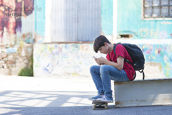 Seitenansicht eines Schülers mit Skateboard  der ein Mobiltelefon benutzt  während er in der Stadt sitzt