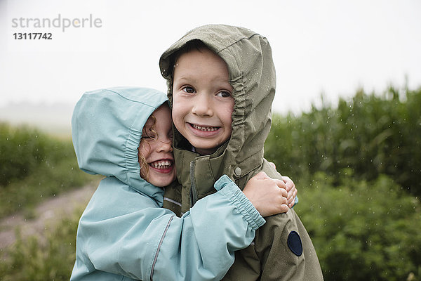 Glückliche Schwester umarmt Bruder  während sie in der Regenzeit gegen Pflanzen steht