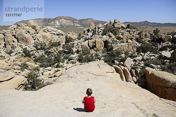 Rückansicht eines Jungen  der Felsformationen betrachtet  während er im Joshua-Tree-Nationalpark gegen den Himmel sitzt