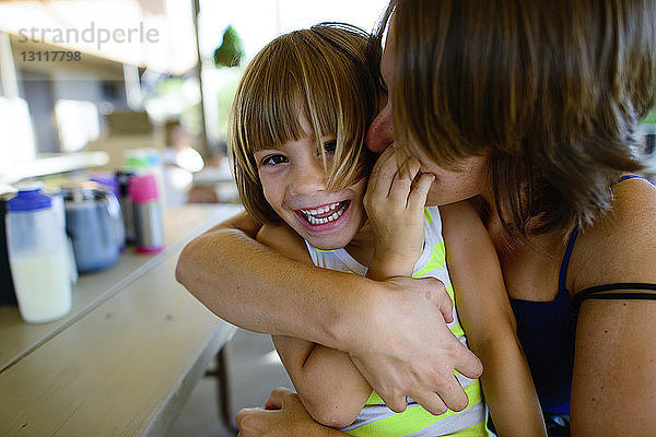 Mutter küsst und umarmt glückliche Tochter  während sie am Tisch im Hof sitzt