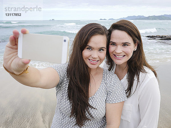Fröhliche Schwestern  die sich mit dem Handy selbstständig machen  während sie am Strand gegen den Himmel stehen