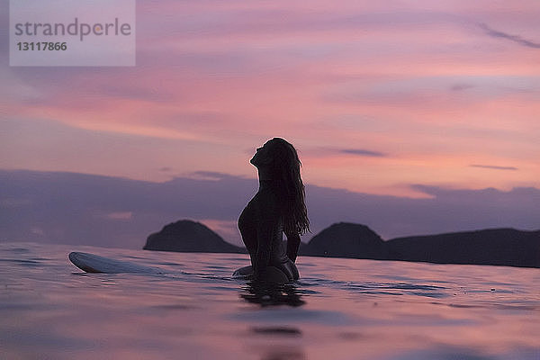 Silhouette einer Frau  die bei Sonnenuntergang auf dem Meer vor bewölktem Himmel surft