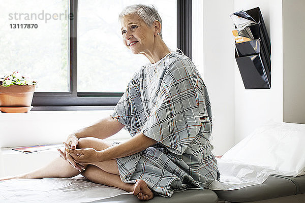Glückliche nachdenkliche Frau schaut weg  während sie in der Klinik auf dem Bett sitzt