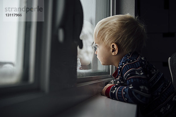 Seitenansicht eines kleinen Jungen  der zu Hause stehend durch ein Fenster schaut