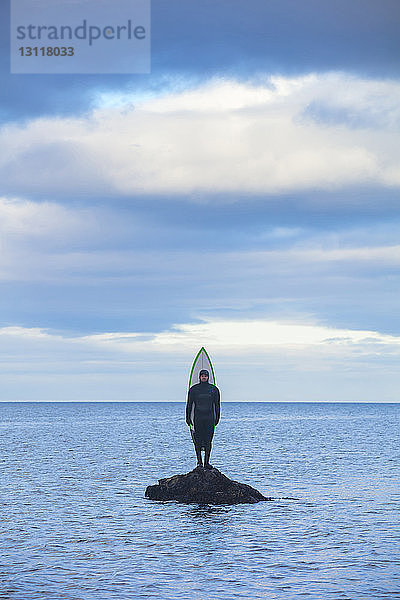 Mann im Neoprenanzug  während er mit einem Surfbrett auf einem Felsen inmitten des Meeres vor bewölktem Himmel steht