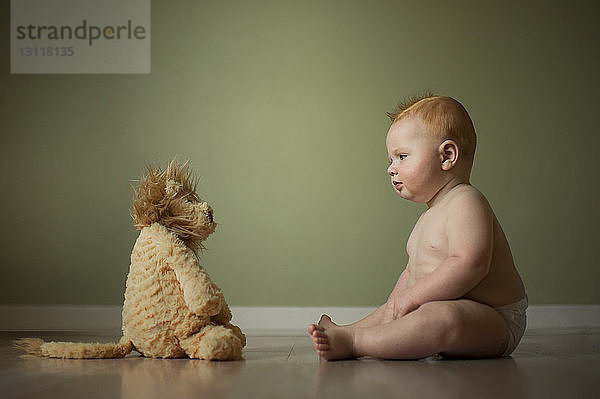 Kleiner Junge ohne Hemd betrachtet Stofftier  während er zu Hause am Boden an der Wand sitzt