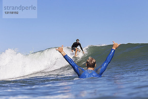 Mann jubelt  während er einen Freund beim Surfen auf dem Meer vor klarem blauen Himmel betrachtet