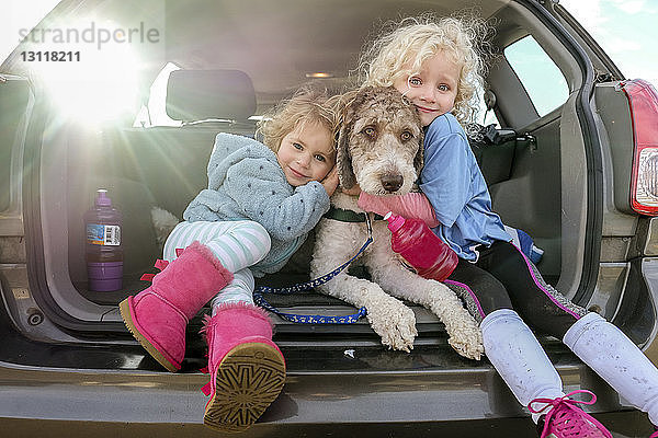 Porträt von lächelnden Schwestern  die einen Hund umarmen  der im Kofferraum eines Autos sitzt