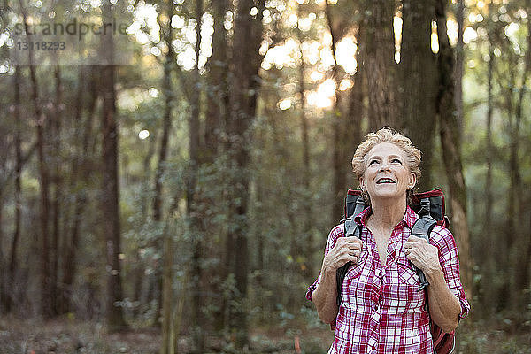 Lächelnde ältere Frau mit Rucksack steht an Bäumen im Wald