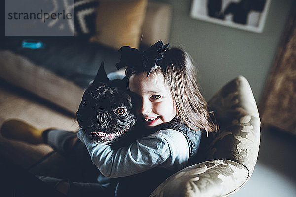 Hochwinkelporträt eines glücklichen Mädchens  das die französische Bulldogge umarmt  während es zu Hause auf dem Sofa sitzt