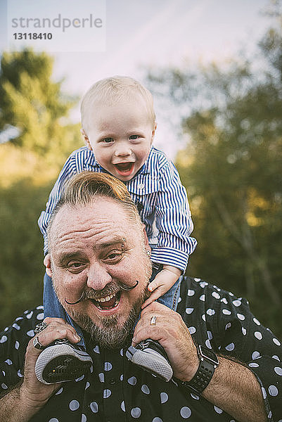 Porträt eines fröhlichen Vaters  der einen süßen Sohn auf den Schultern trägt  im Park