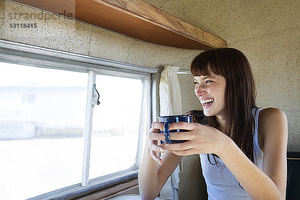 Fröhliche Frau trinkt Kaffee  während sie im Wohnmobil am Fenster sitzt