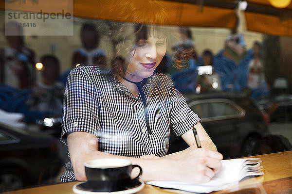 Frau schreibt auf Buch  während sie im Café sitzt und durch ein Fenster gesehen wird