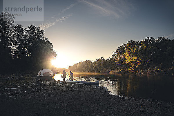 Männliche Freunde gehen bei Sonnenuntergang mit dem Boot am Seeufer gegen den Himmel