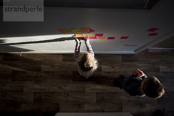 Hochwinkelansicht von Geschwistern  die zu Hause Haftnotizen an die Wand kleben