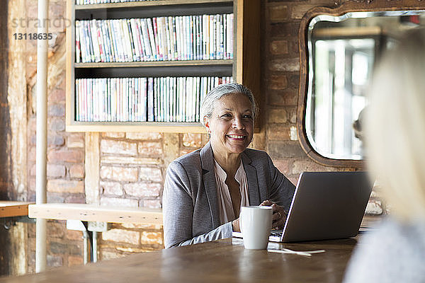 Lächelnde Frau mit Laptop  während sie im Café am Bücherregal sitzt