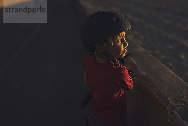 Seitenansicht eines Jungen mit Fahrradhelm  der bei Sonnenuntergang an der Stützmauer steht