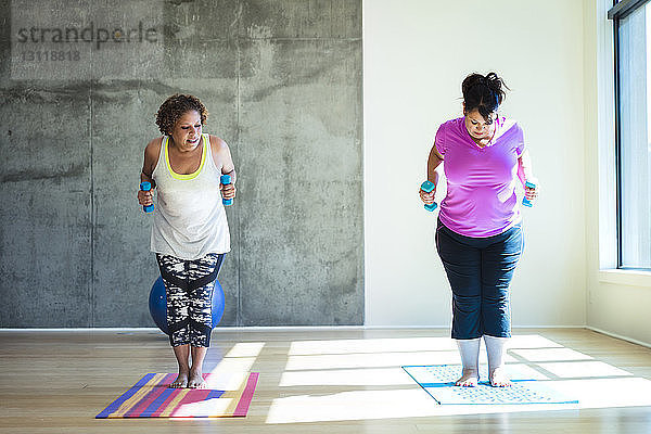 Frau sieht Freundin an  die auf Übungsmatte steht  während sie im Yoga-Studio Hanteln gegen die Wand hält