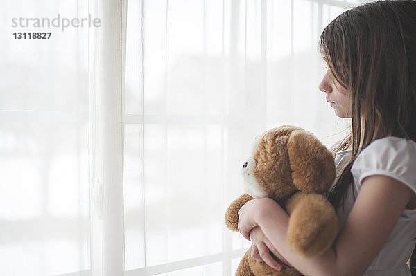 Mädchen hält Teddybär und schaut zu Hause durchs Fenster