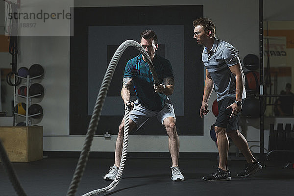 Trainer betrachtet männlichen Kunden beim Training mit Seilen im Fitnessstudio