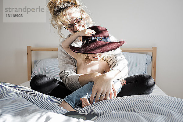 Glückliche Mutter mit hemdloser Tochter mit Hut  die zu Hause auf dem Bett sitzt