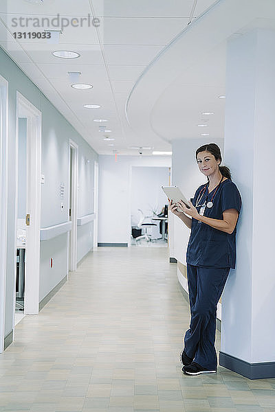 Porträt einer Ärztin  die einen Tablet-Computer benutzt  während sie sich im Krankenhaus an eine architektonische Säule lehnt