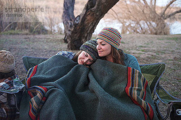 Liebevolle Freundinnen mit Decke zugedeckt auf dem Zeltplatz sitzend