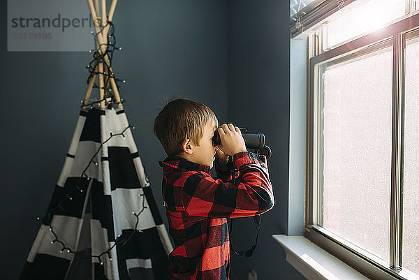 Seitenansicht eines Jungen  der zu Hause am Fenster durch ein Fernglas schaut