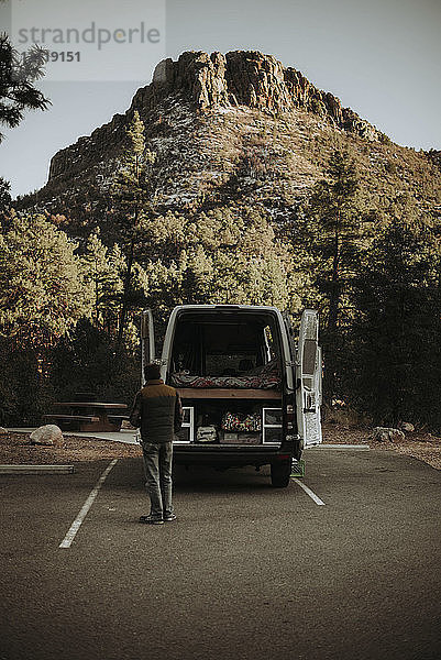 Rückansicht eines Mannes  der an einem Wohnmobil auf einer Straße gegen einen Berg im Wald steht