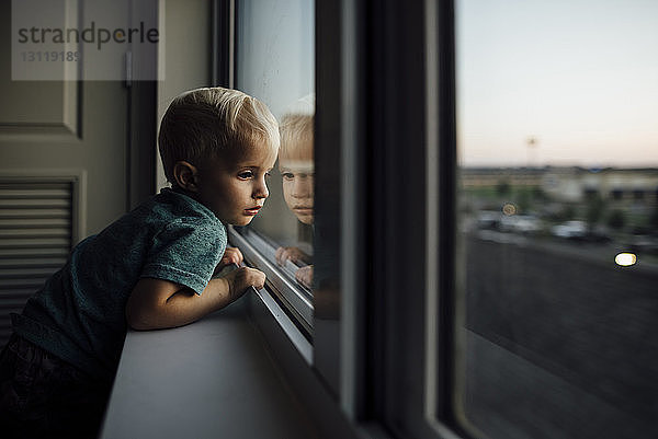 Seitenansicht eines kleinen Jungen  der zu Hause stehend durch ein Fenster schaut