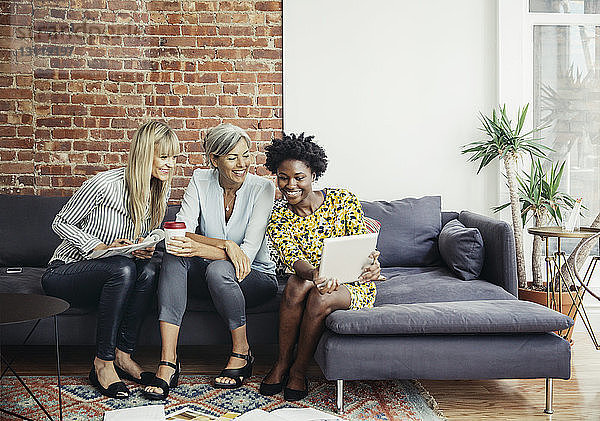 Fröhliche Geschäftsfrauen  die im Kreativbüro auf dem Sofa sitzend einen Tablet-Computer benutzen