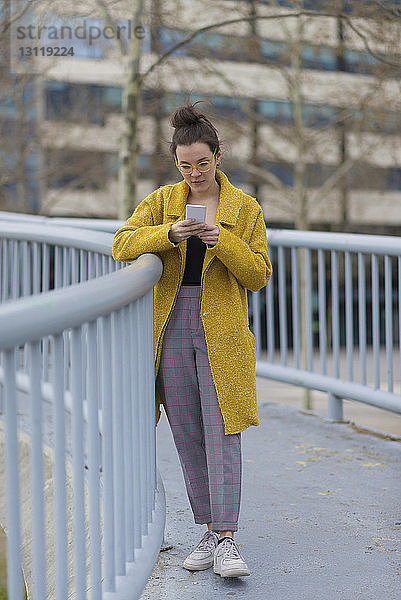 Selbstbewusste Geschäftsfrau benutzt Smartphone  während sie auf einer Fußgängerbrücke gegen Gebäude in der Stadt steht