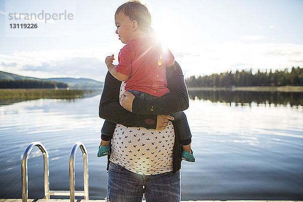 Schwangere Mutter trägt Sohn am See bei sonnigem Wetter