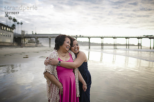 Glückliche Frau umarmt Mutter  während sie am Strand vor bewölktem Himmel steht