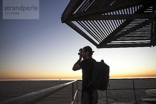 Seitenansicht eines Rucksacktouristen  der bei Sonnenuntergang mit der Kamera gegen den klaren Himmel am Strand fotografiert